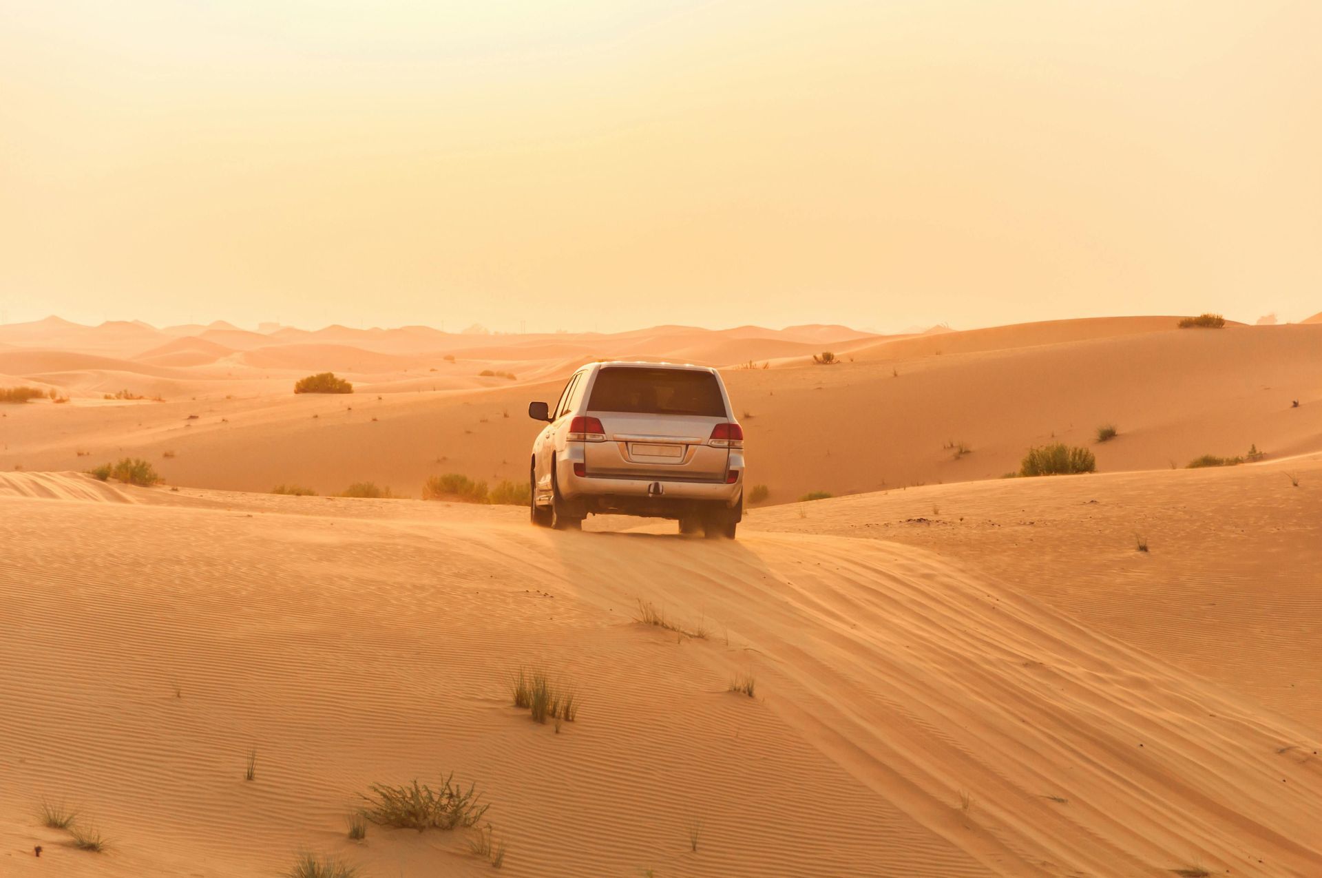 Un SUV argentato percorre vaste dune di sabbia in un paesaggio desertico durante un tramonto dorato, sollevando sabbia.