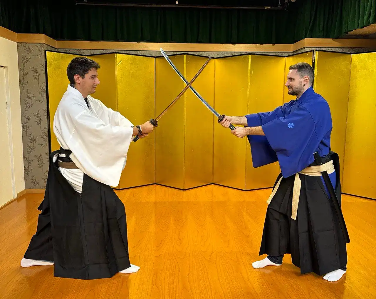 Due uomini in abiti tradizionali giapponesi duellano con le katane, le spade incrociate davanti a un paravento dorato.