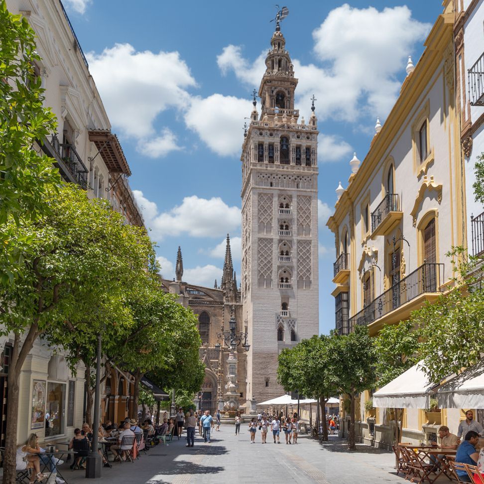 An ornate bell tower stands at the end of a sunny European street lined with trees, cafes, and strolling people under a blue sky.