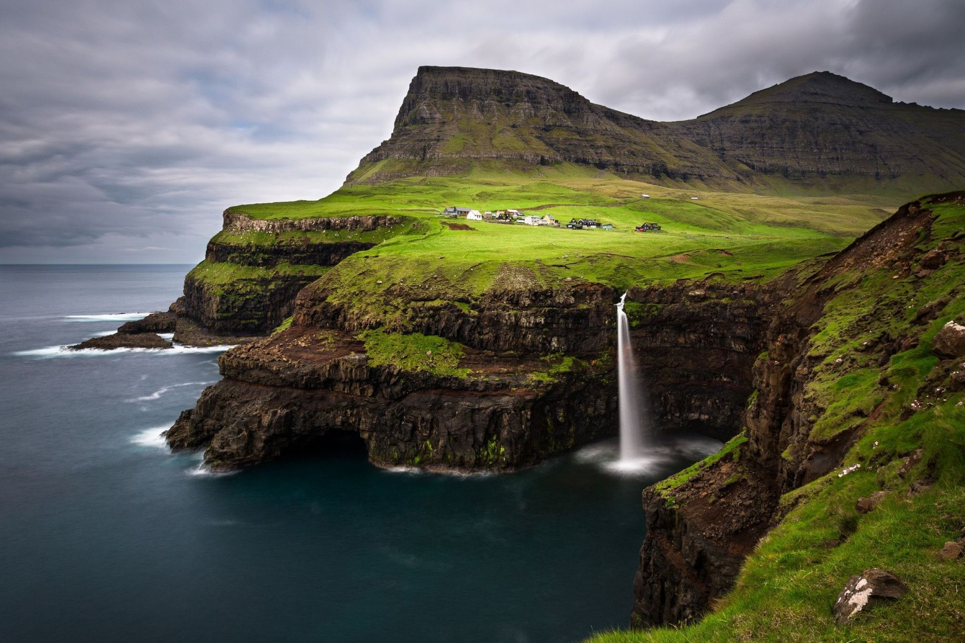 Una cascata si riversa da una verde scogliera erbosa direttamente nell'oceano, con un piccolo villaggio incastonato sulla collina sullo sfondo.