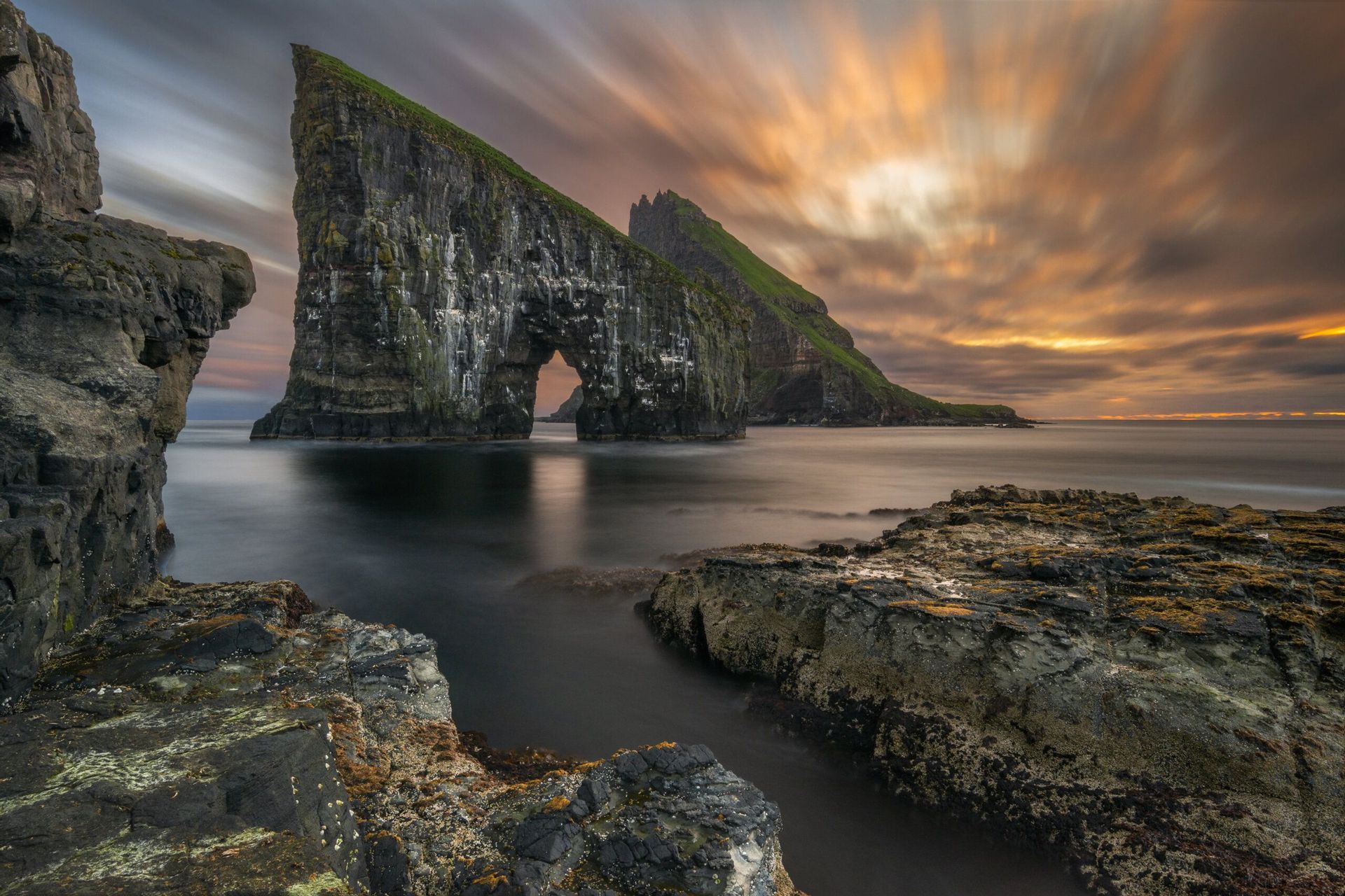Scatto a lunga esposizione di due grandi faraglioni, uno con un arco naturale, su una costa rocciosa sotto un drammatico cielo al tramonto.