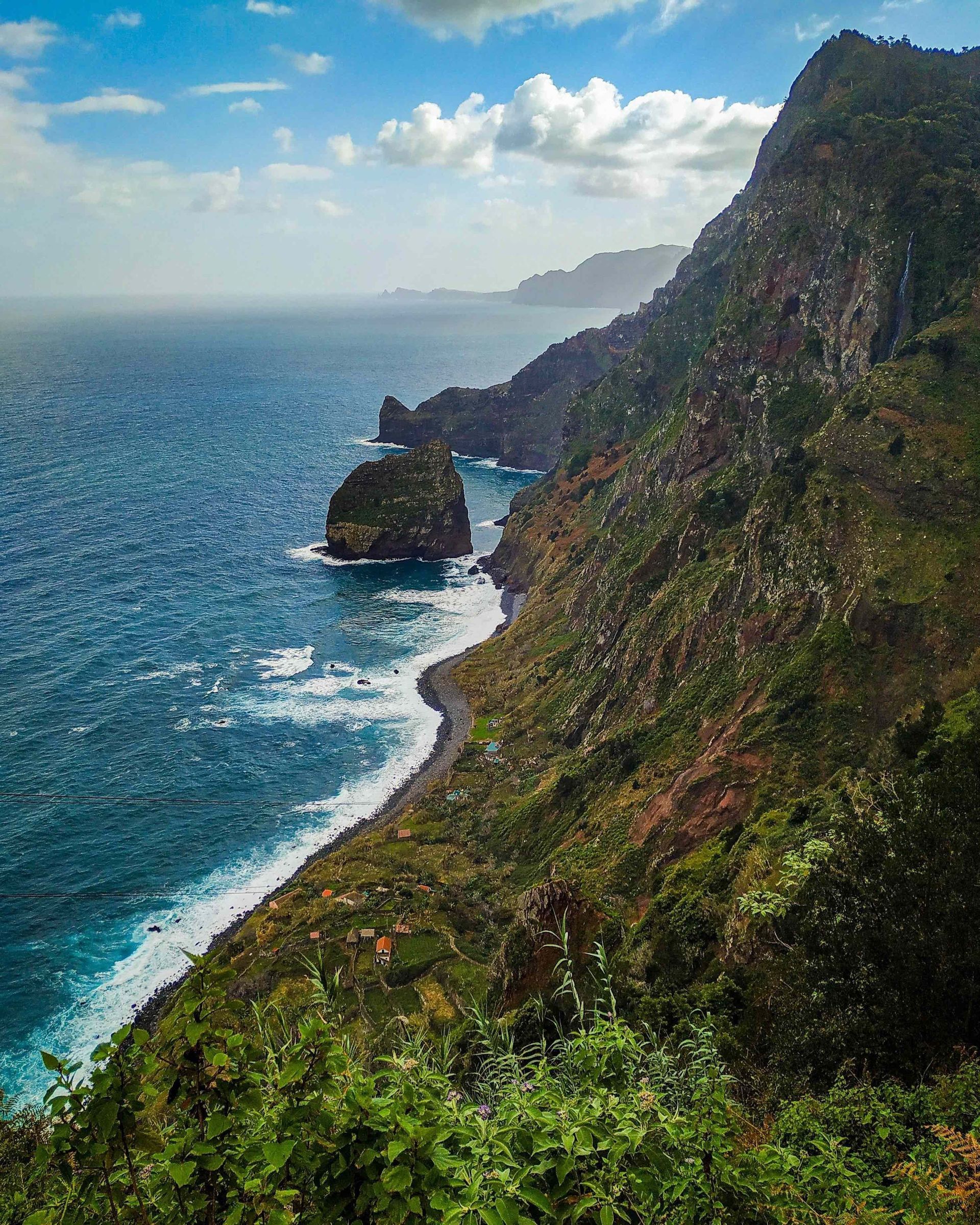 Una ripida scogliera verde incontra l'oceano blu lungo una costa frastagliata, con onde che lambiscono una piccola spiaggia e un villaggio sottostante.