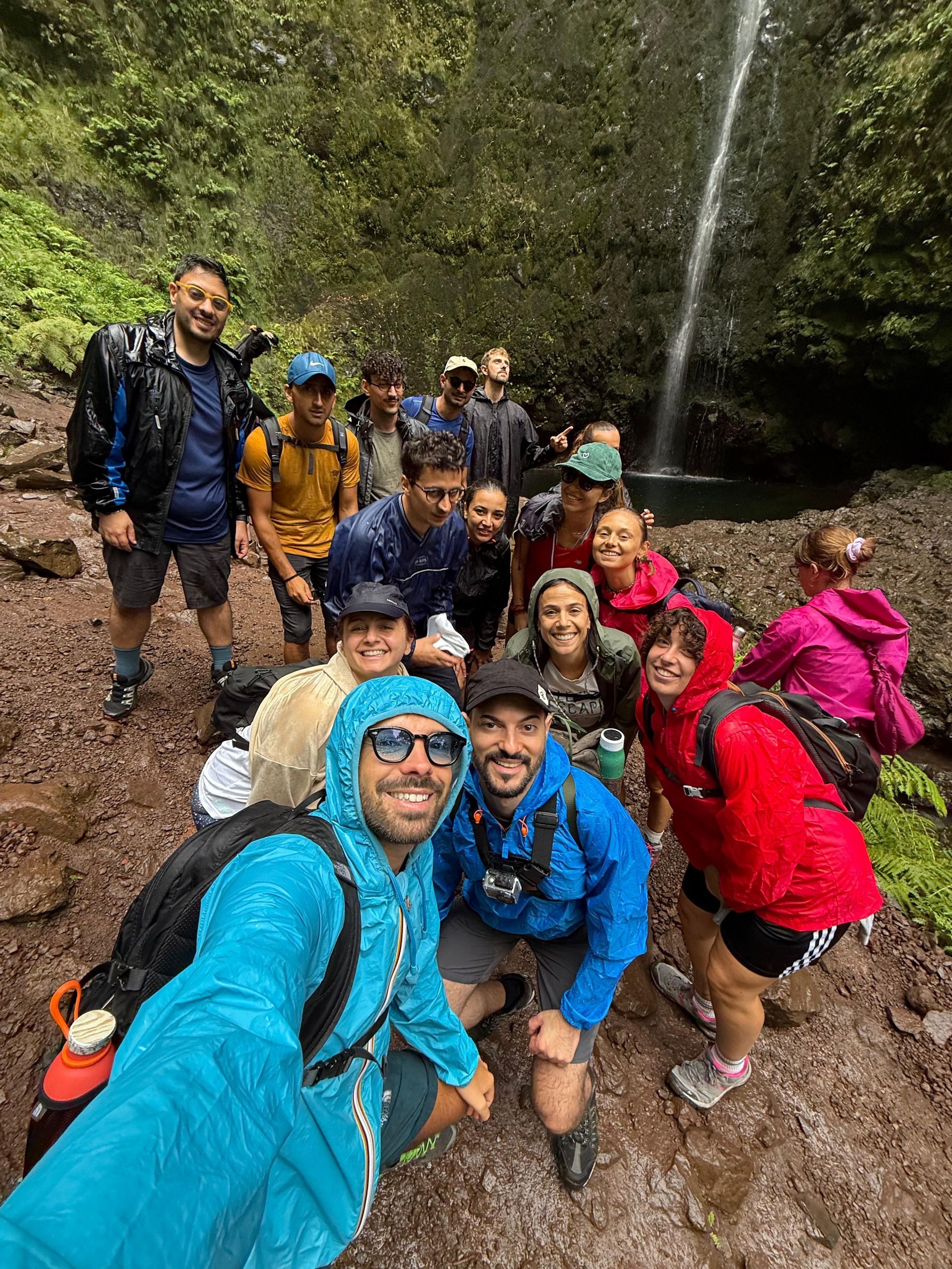 Un gruppo WeRoad in impermeabili colorati posa per un selfie davanti a un'alta cascata in una foresta lussureggiante.