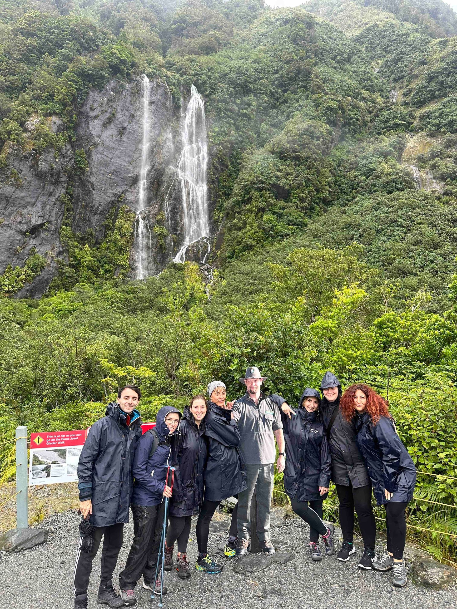 A WeRoad group trip wearing raincoats poses for a photo in front of a waterfall on a lush green mountainside.