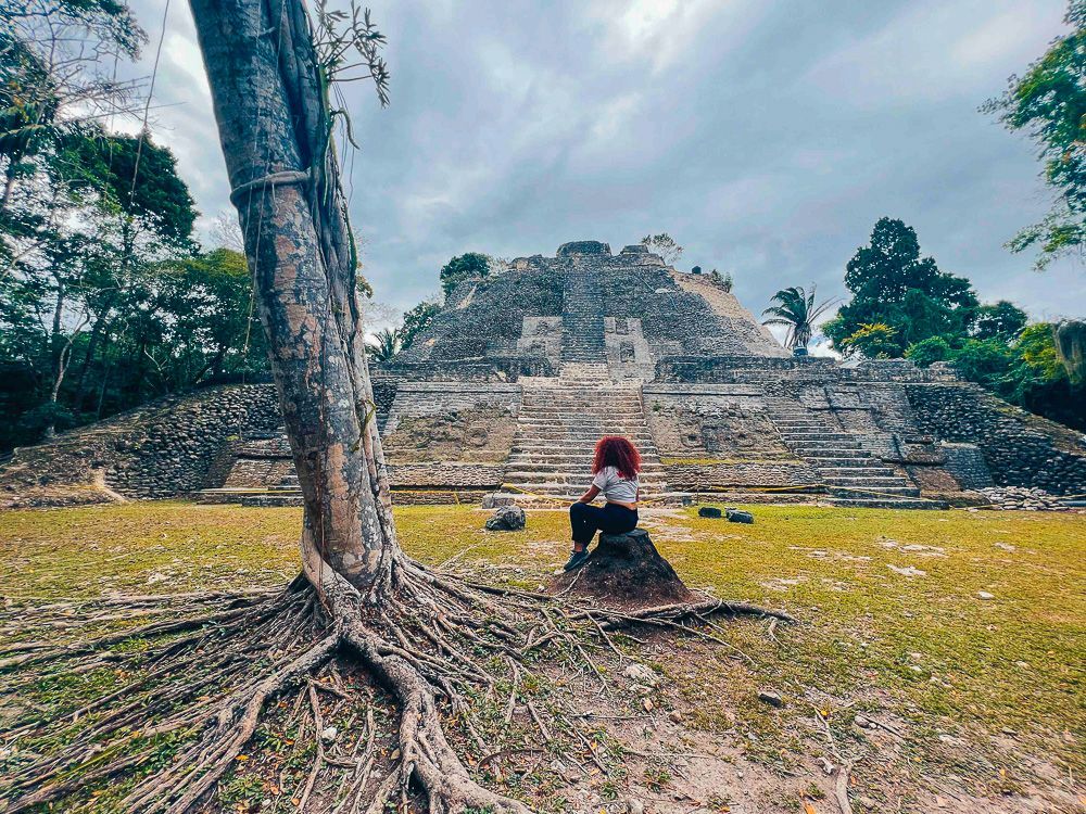 Una persona dai capelli rossi siede su una roccia ammirando un'antica piramide di pietra, con le radici di un grande albero in primo piano.