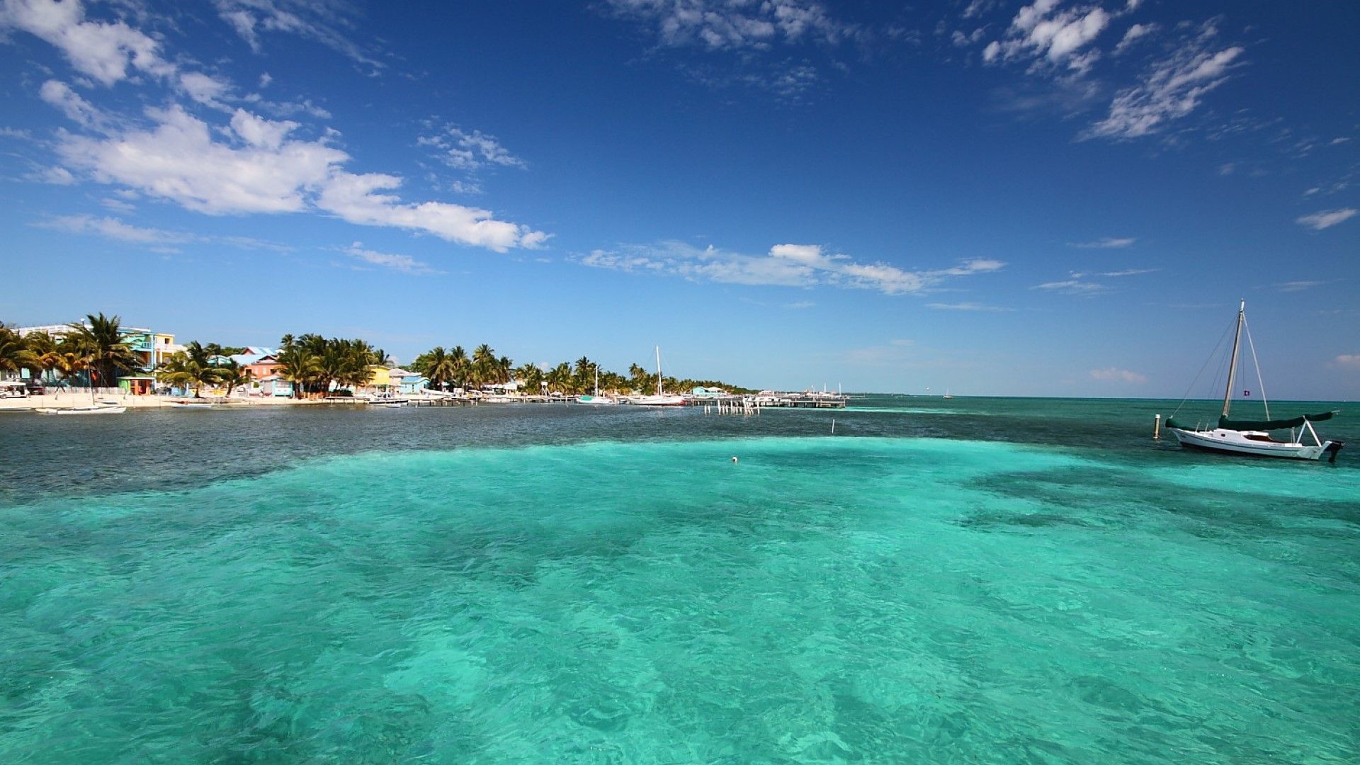 Una vista sull'acqua limpida e turchese che mostra una costa tropicale con palme, edifici colorati e una barca a vela bianca ancorata.