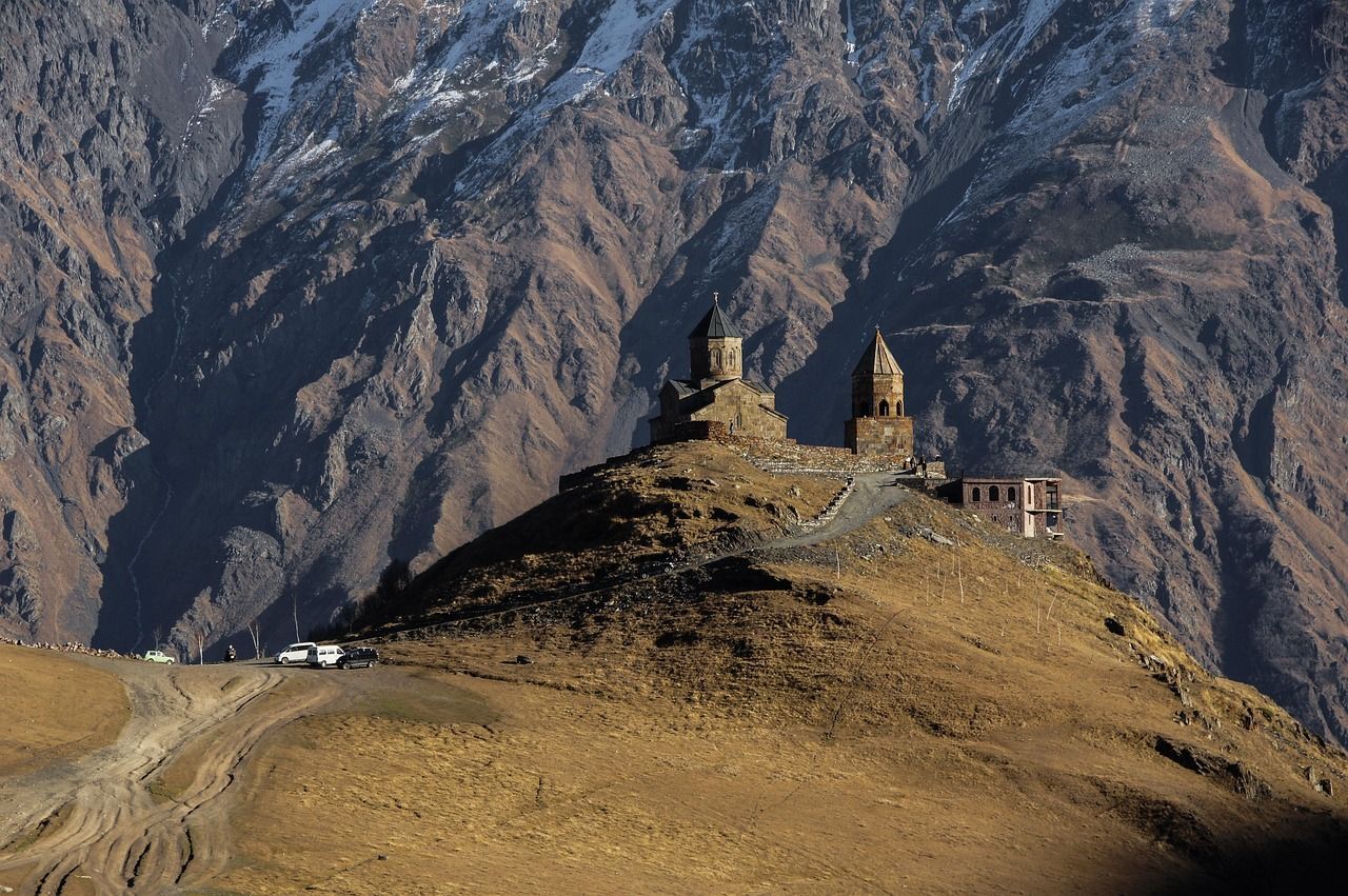Una chiesa e un campanile in pietra si ergono su una collina erbosa, con una massiccia e aspra catena montuosa sullo sfondo.