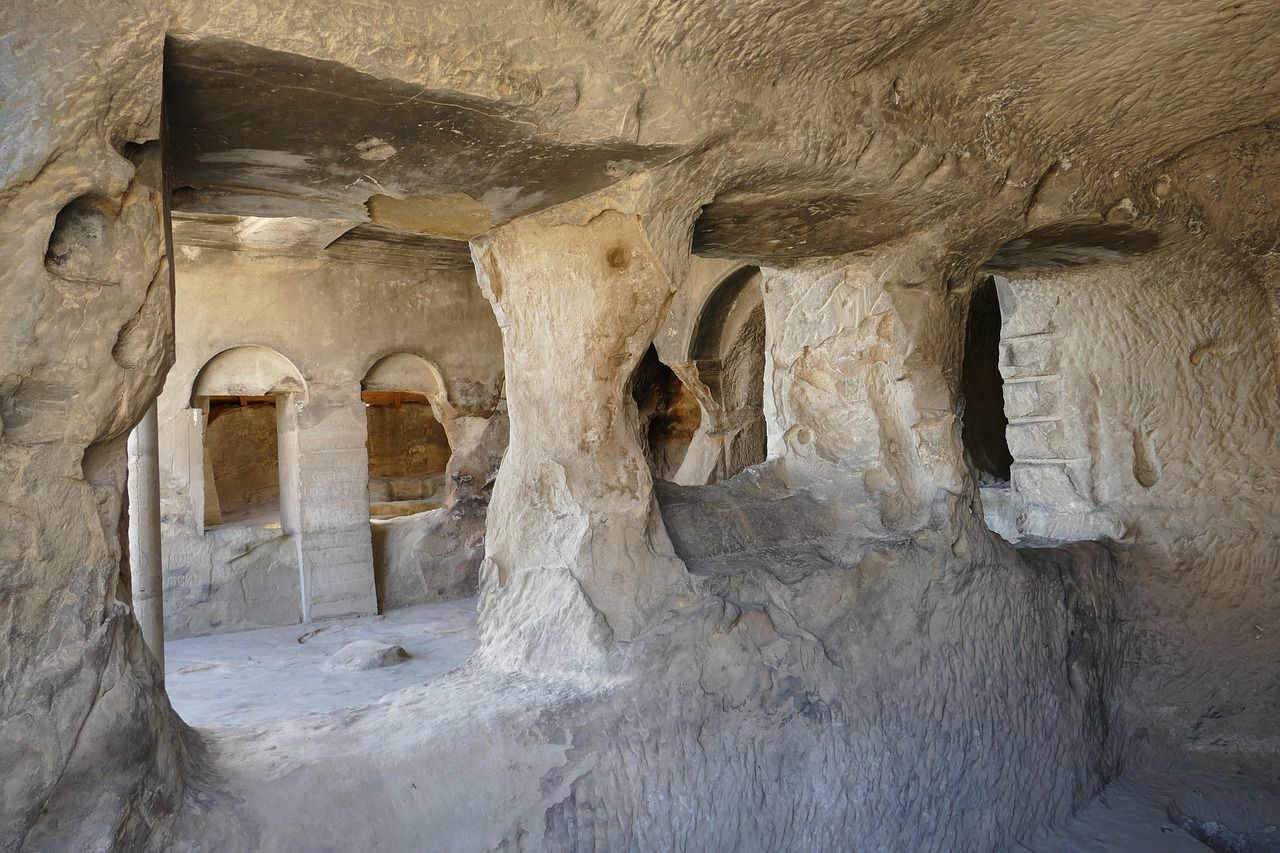 L'interno di un'abitazione rupestre scavata a mano con pilastri in pietra e finestre ad arco sotto un soffitto di roccia grezza.
