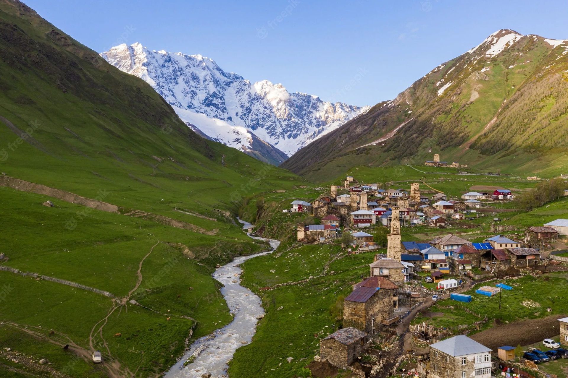Una vista aerea di un piccolo villaggio con torri in pietra in una valle verde, situato vicino a un fiume con montagne innevate sullo sfondo.