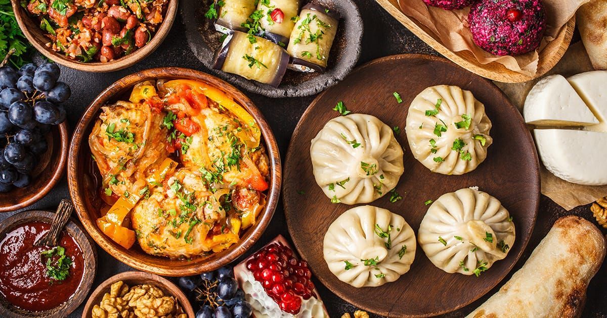 Una vista dall'alto di un ricco assortimento di piatti tradizionali, tra cui ravioli, stufati, insalate, formaggi e frutta fresca, disposti su una superficie scura.