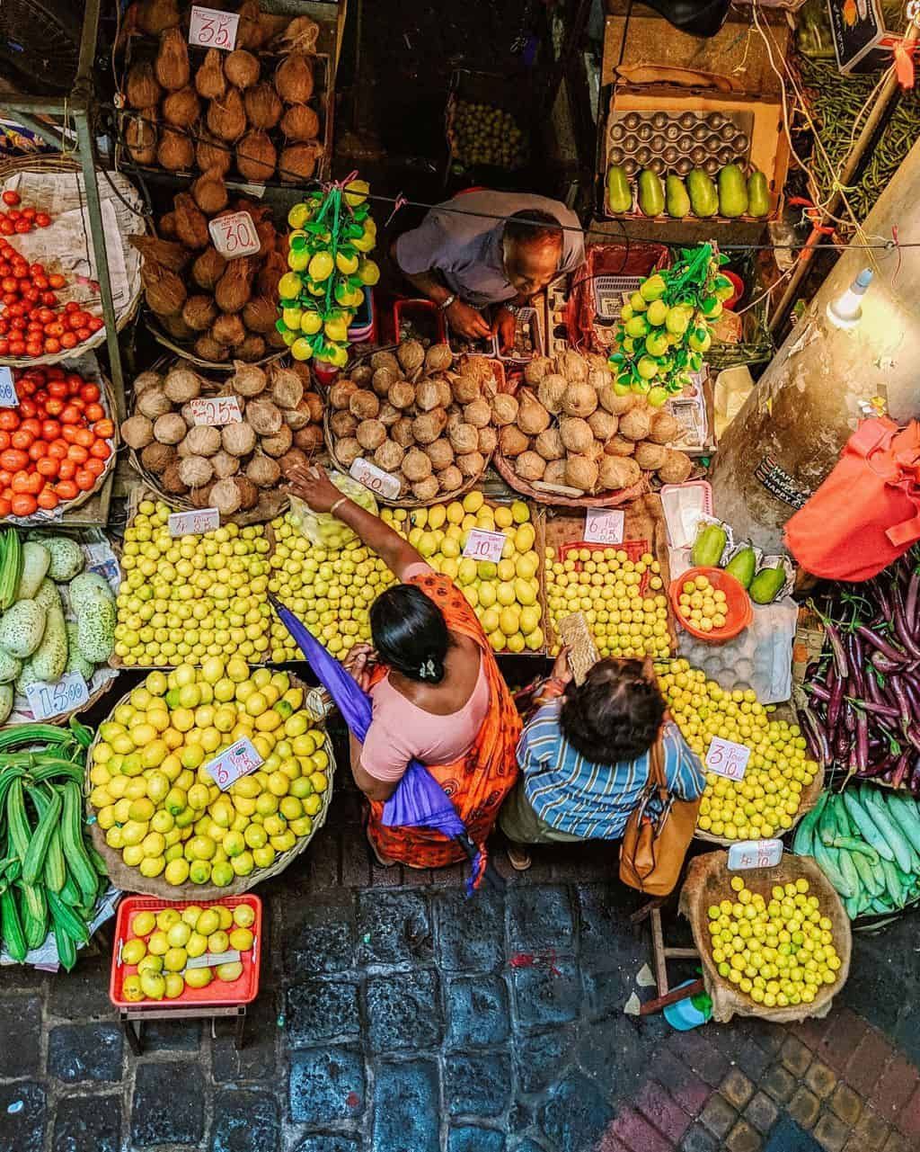 Vista dall'alto di due donne che acquistano limoni e noci di cocco da un venditore in un affollato mercato all'aperto.
