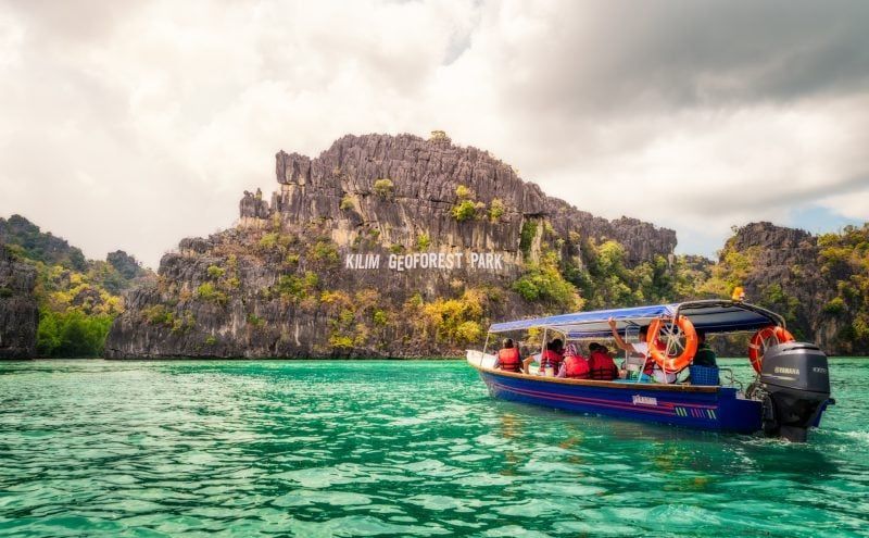 Eine WeRoad-Gruppenreise auf einem Boot segelt auf türkisfarbenem Wasser und nähert sich dem Schild des Kilim Geoforest Parks auf einer felsigen Insel.