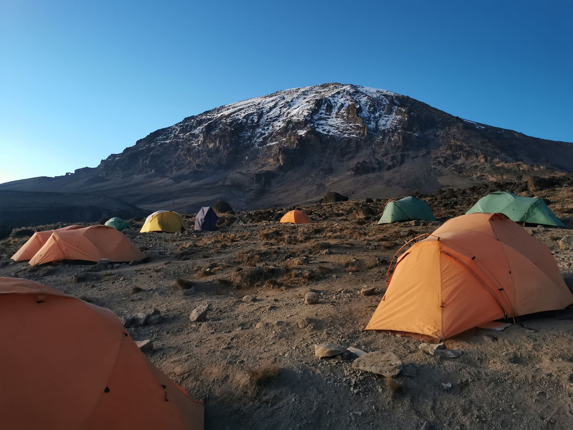 Tende colorate punteggiano un campeggio roccioso ai piedi di una montagna innevata sotto un cielo azzurro e limpido.