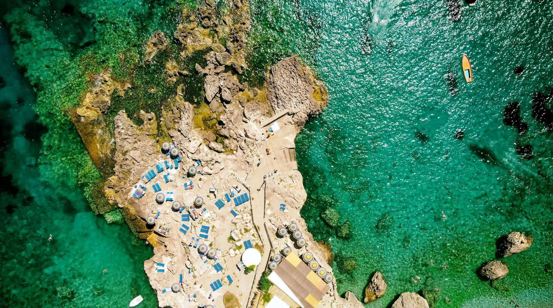 An aerial top-down view of a rocky coastline with blue sun loungers and umbrellas next to vibrant turquoise water.