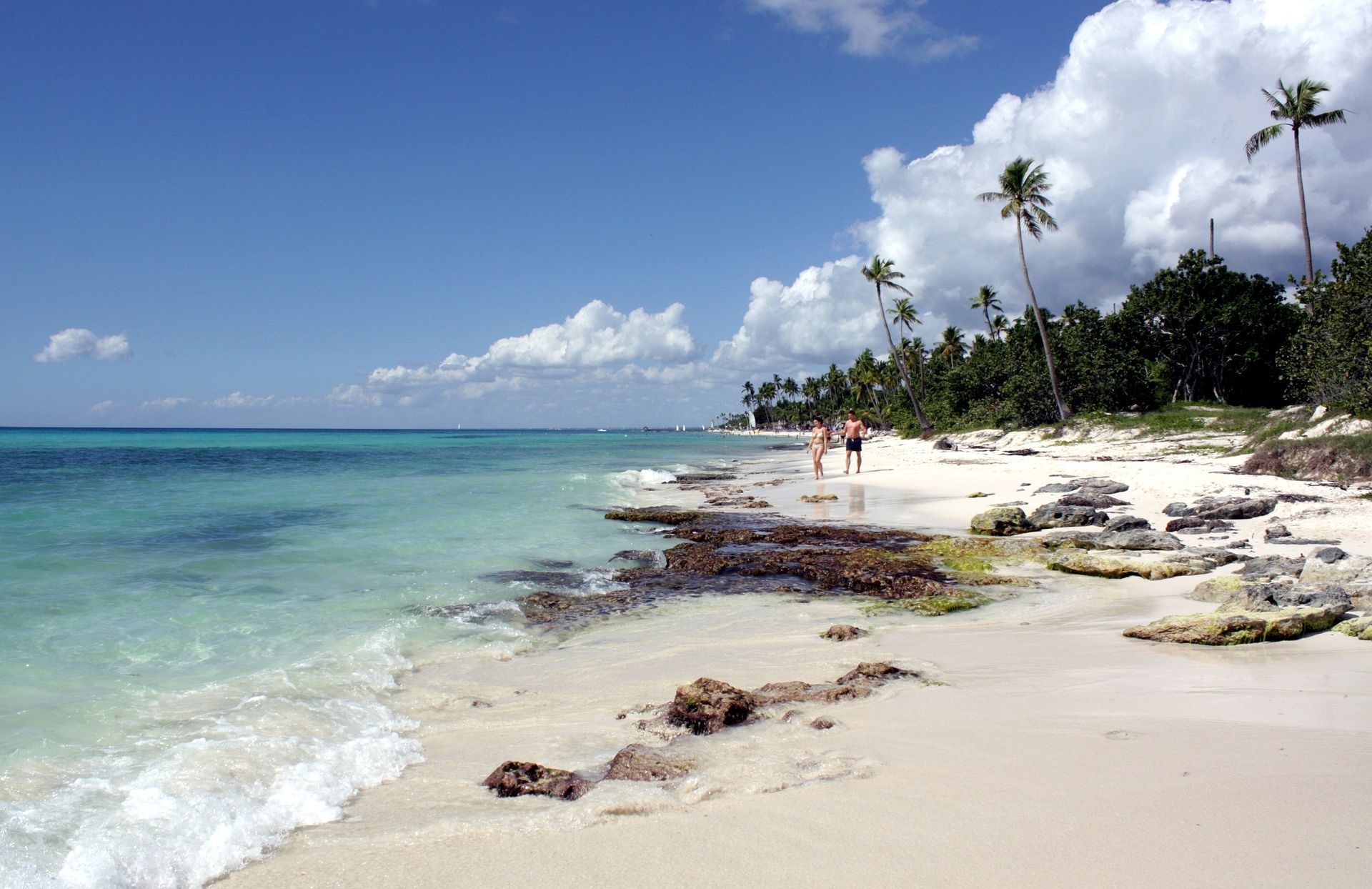 Two people walk on a tropical beach with white sand and turquoise water, lined with tall palm trees.