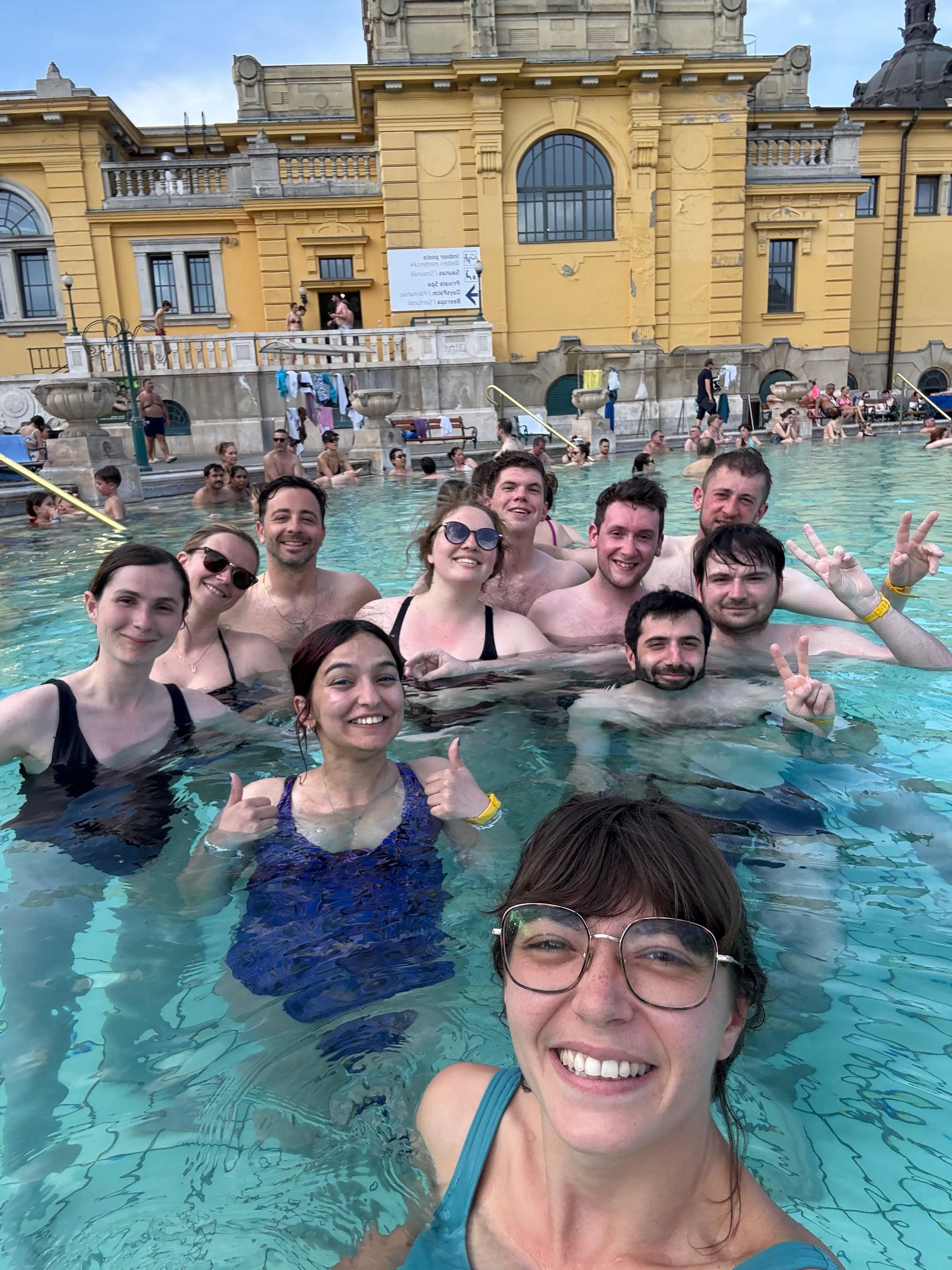 Eine WeRoad-Gruppe macht lächelnd ein Selfie in einem Freibad vor einem großen, gelben historischen Gebäude.