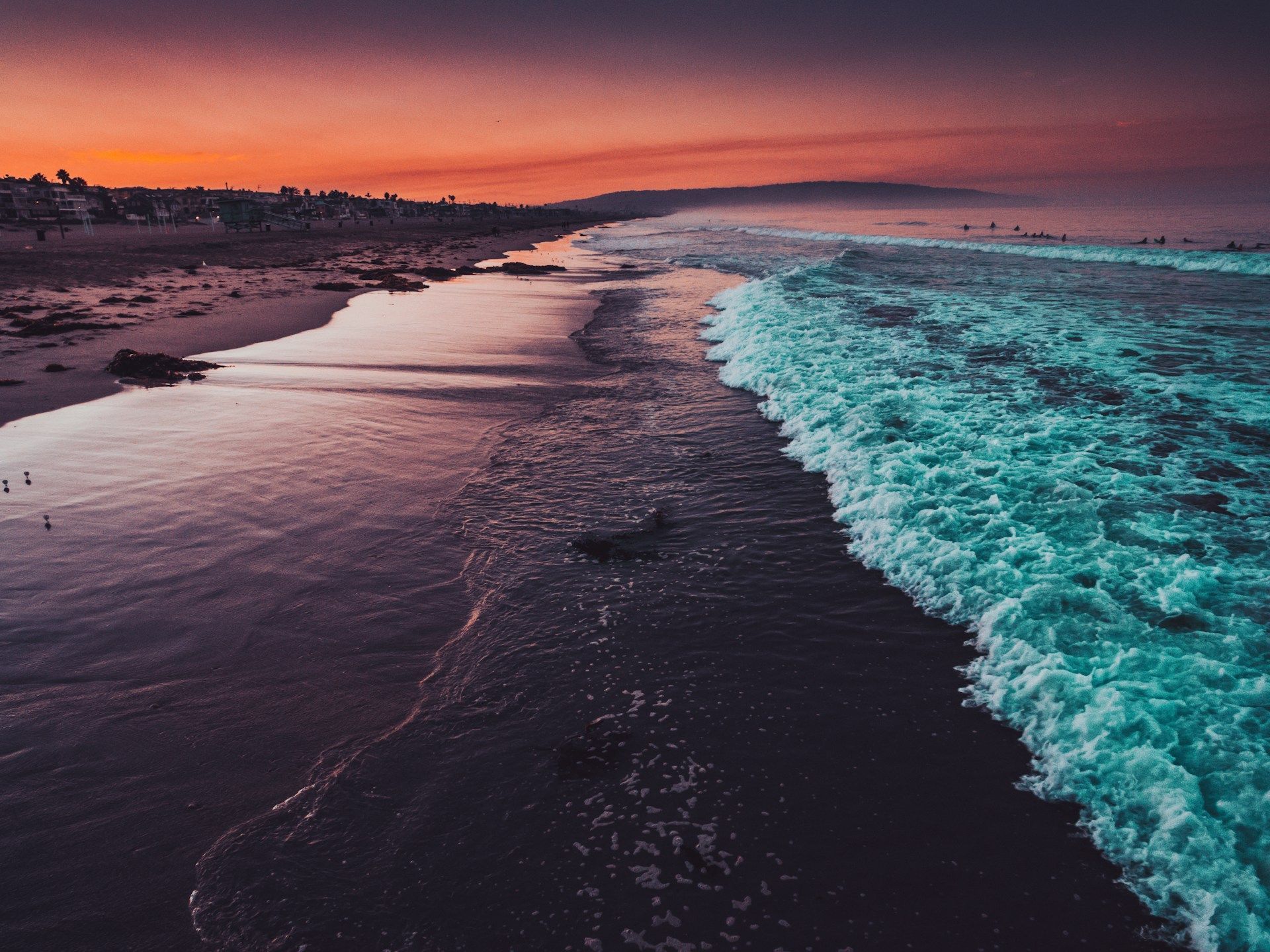 Turquoise waves roll onto a dark sand beach, reflecting the vibrant orange and purple colors of the sunset sky.
