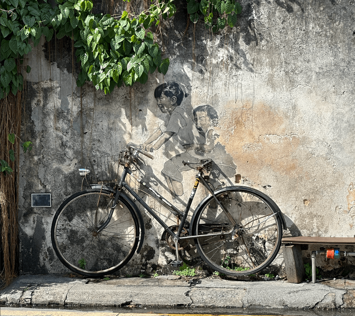 Una vera bicicletta è parcheggiata contro un muro logoro con un murale di street art raffigurante due bambini che la cavalcano.