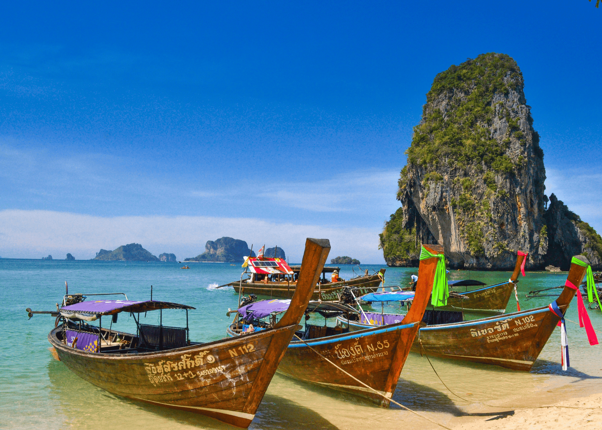 Barche a coda lunga thailandesi ormeggiate su una spiaggia sabbiosa in acque turchesi, con faraglioni calcarei sullo sfondo sotto un cielo azzurro.