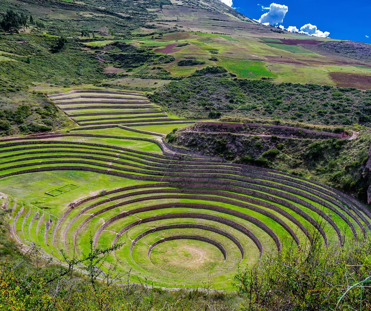 Concentric circular agricultural terraces with stone walls built into a lush green hillside under a blue sky.