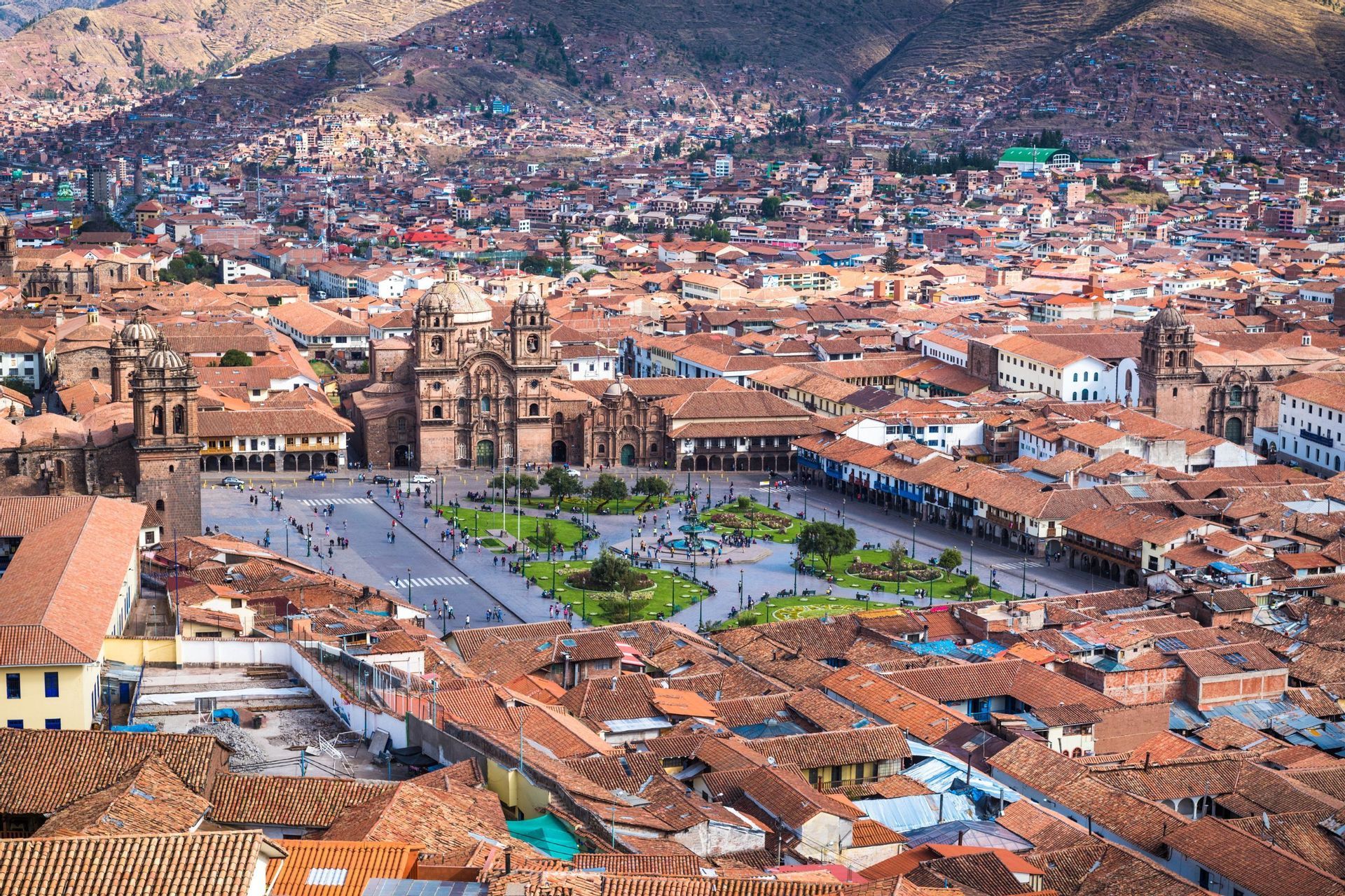 An aerial view of a bustling city square surrounded by historic buildings with terracotta roofs, with mountains visible in the background.