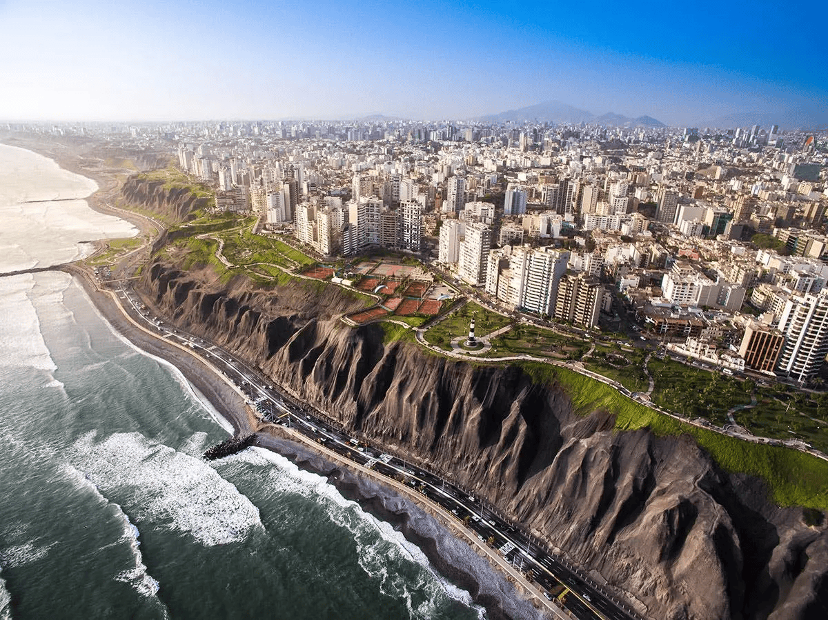 An aerial view of a coastal city built on steep cliffs, with a highway below and waves crashing on the beach.