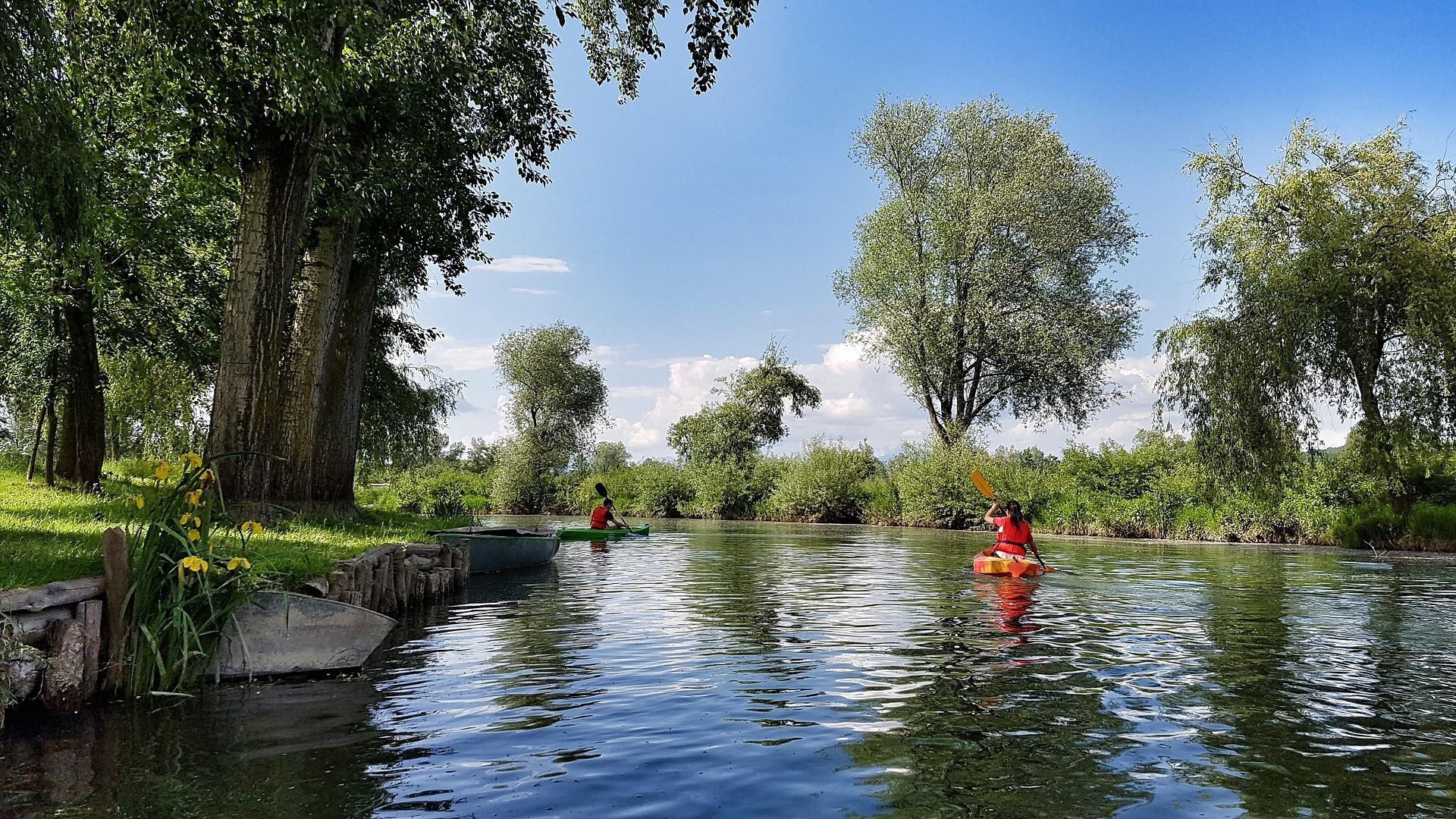 Due persone di un gruppo WeRoad in kayak su un fiume tranquillo, con alberi rigogliosi sulle sponde sotto un cielo azzurro.