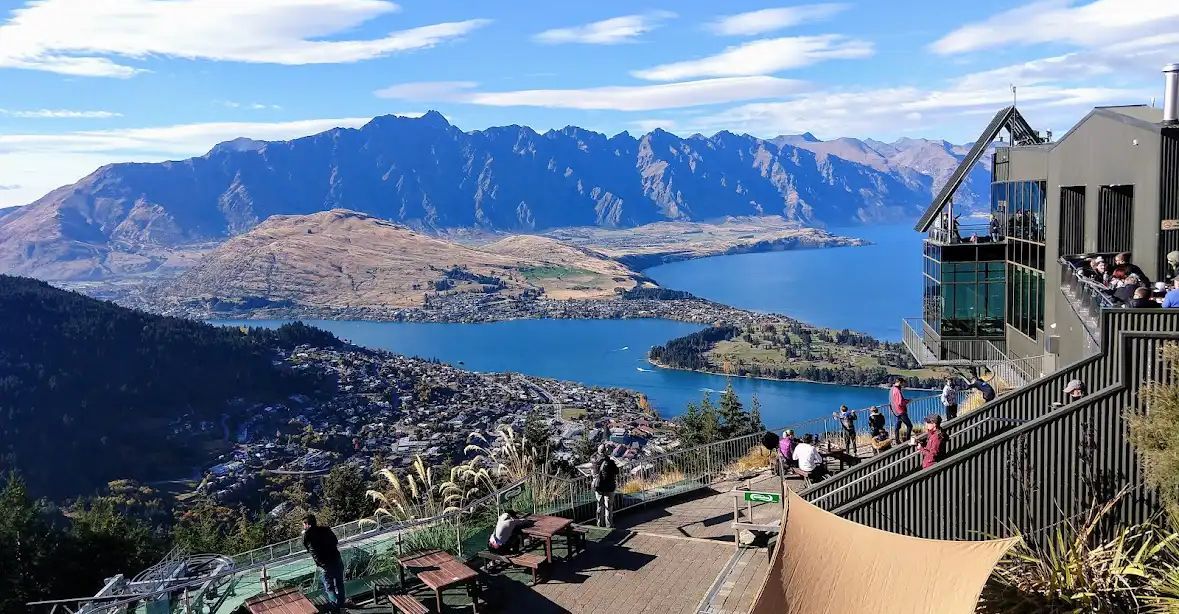 A group of people on a modern viewing deck looking out over a city, a large blue lake, and a distant mountain range.