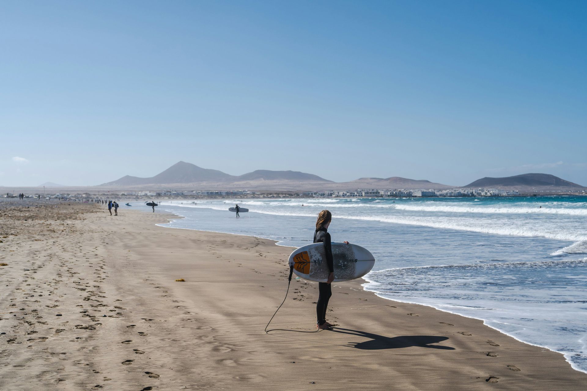 Un surfista in muta tiene una tavola da surf su una spiaggia sabbiosa, guardando l'oceano con montagne distanti sotto un cielo sereno.