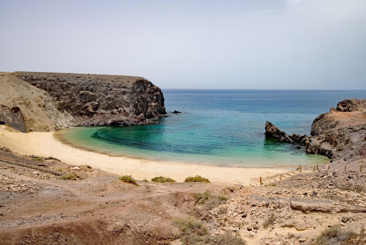 Panoramica da una scogliera rocciosa su una spiaggia sabbiosa appartata, incastonata in una caletta con acque turchesi e cristalline, sotto un cielo leggermente velato.