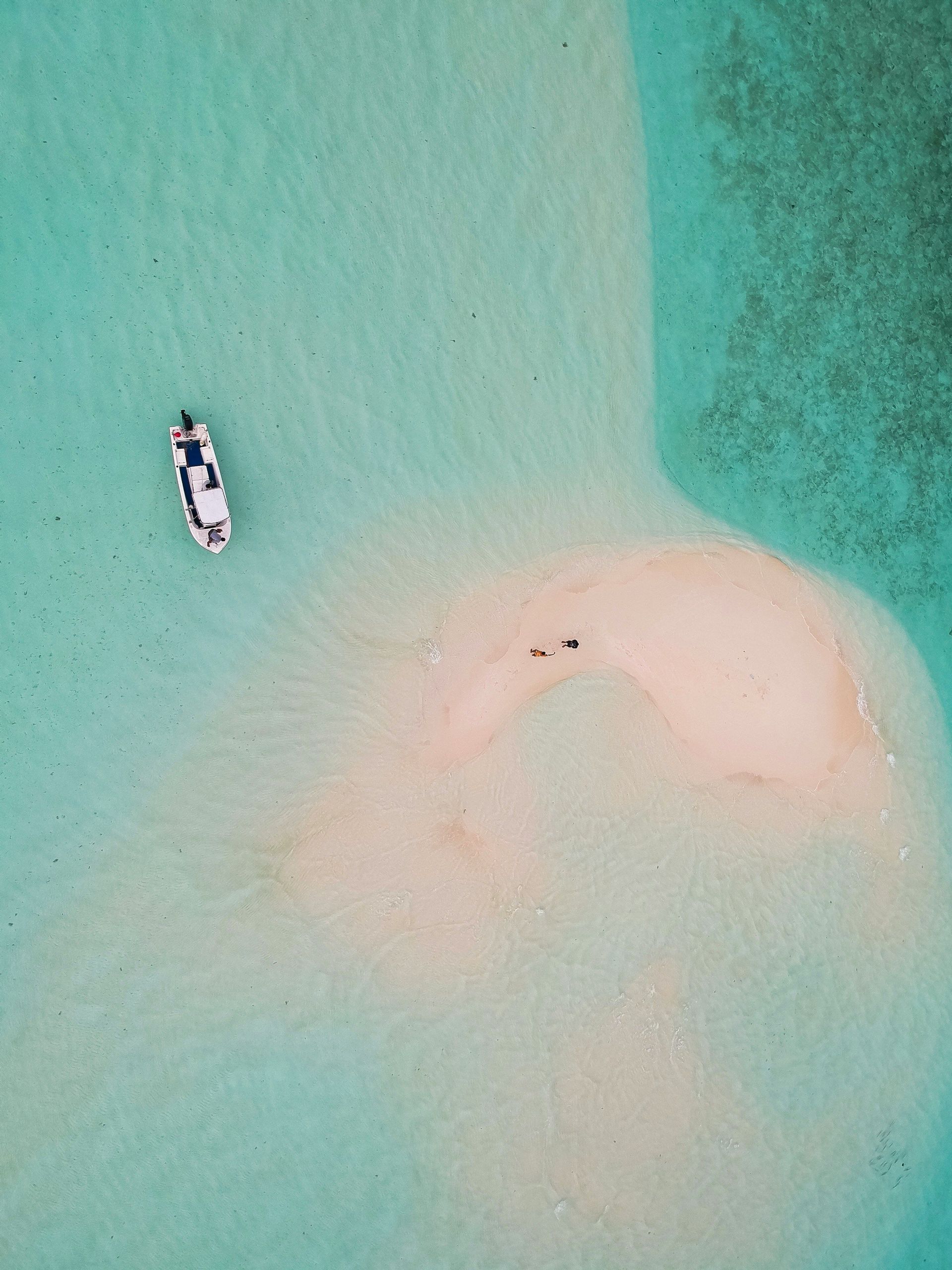 Vista aerea di una piccola barca bianca che galleggia in acqua turchese cristallina, vicino a un banco di sabbia con due persone.