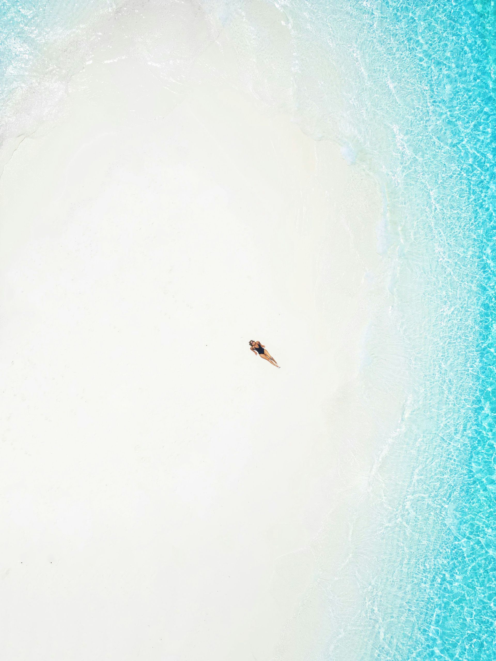 Vista aerea di una persona in costume da bagno sdraiata su una spiaggia di sabbia bianca accanto ad acqua turchese cristallina.
