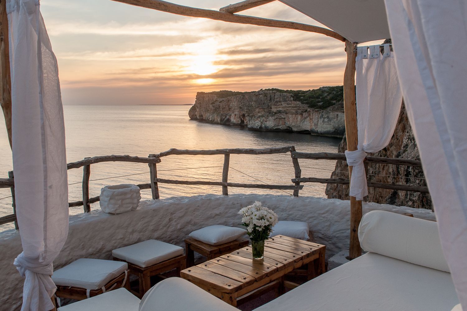 Una terrazza a picco sulla scogliera con mobili in legno e fiori bianchi si affaccia sul mare calmo e una costa rocciosa al tramonto.