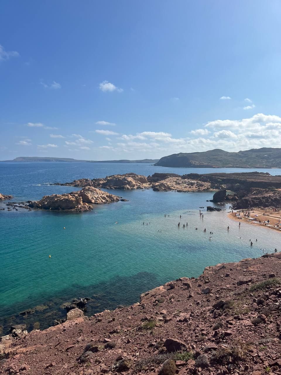 A high-angle view of a rocky cove with turquoise water, where people are swimming and relaxing on a small sandy beach.