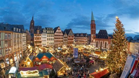 Una vista aérea de un animado mercado de Navidad en una plaza histórica al anochecer, con un gran árbol iluminado y edificios tradicionales.