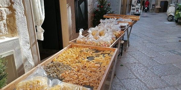 Pasta fresca e multicolore esposta in vassoi di legno e sacchetti su tavoli lungo una stretta via di ciottoli.