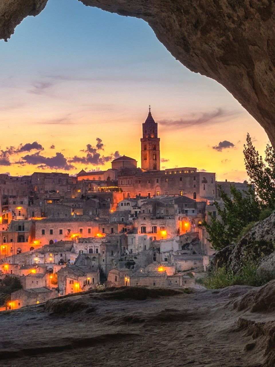 Un'antica città collinare con un campanile imponente, illuminata da luci calde al tramonto e vista dall'interno di una grotta.