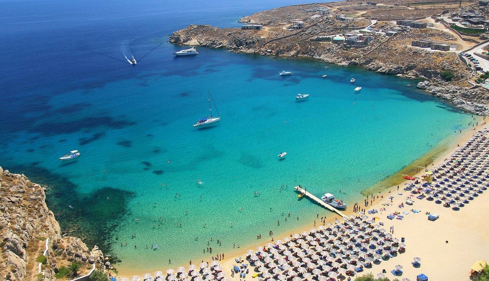 Vista aerea di una spiaggia sabbiosa affollata con ombrelloni, accanto a una baia turchese con bagnanti e barche.