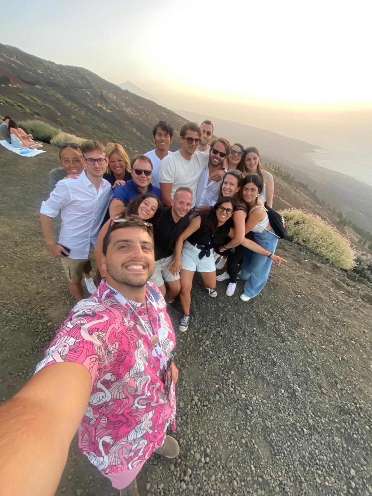 Un voyage de groupe WeRoad souriant pour un selfie pris par un homme en chemise rose sur une pente volcanique au coucher du soleil.