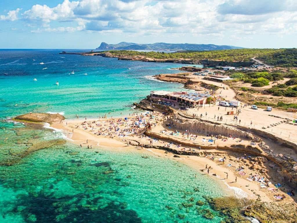 Veduta aerea di una caletta affollata con persone che nuotano e prendono il sole in acque limpide e turchesi vicino a una costa rocciosa in una giornata di sole.