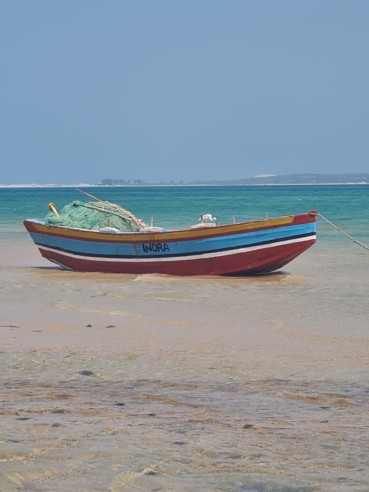 Ein farbenfrohes Fischerboot namens Inora liegt im seichten türkisfarbenen Wasser an einem Sandstrand unter klarem blauem Himmel.