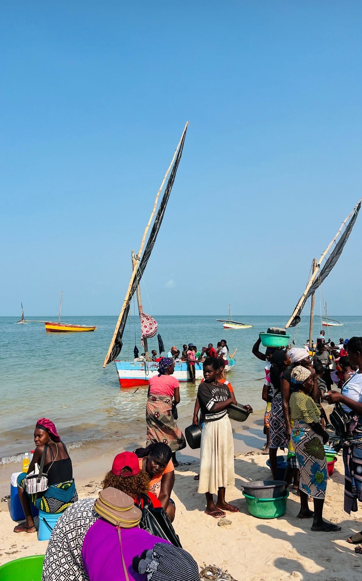 Un gruppo di gente del posto si è riunito su una spiaggia sabbiosa accanto a barche a vela tradizionali sull'acqua sotto un cielo blu chiaro.