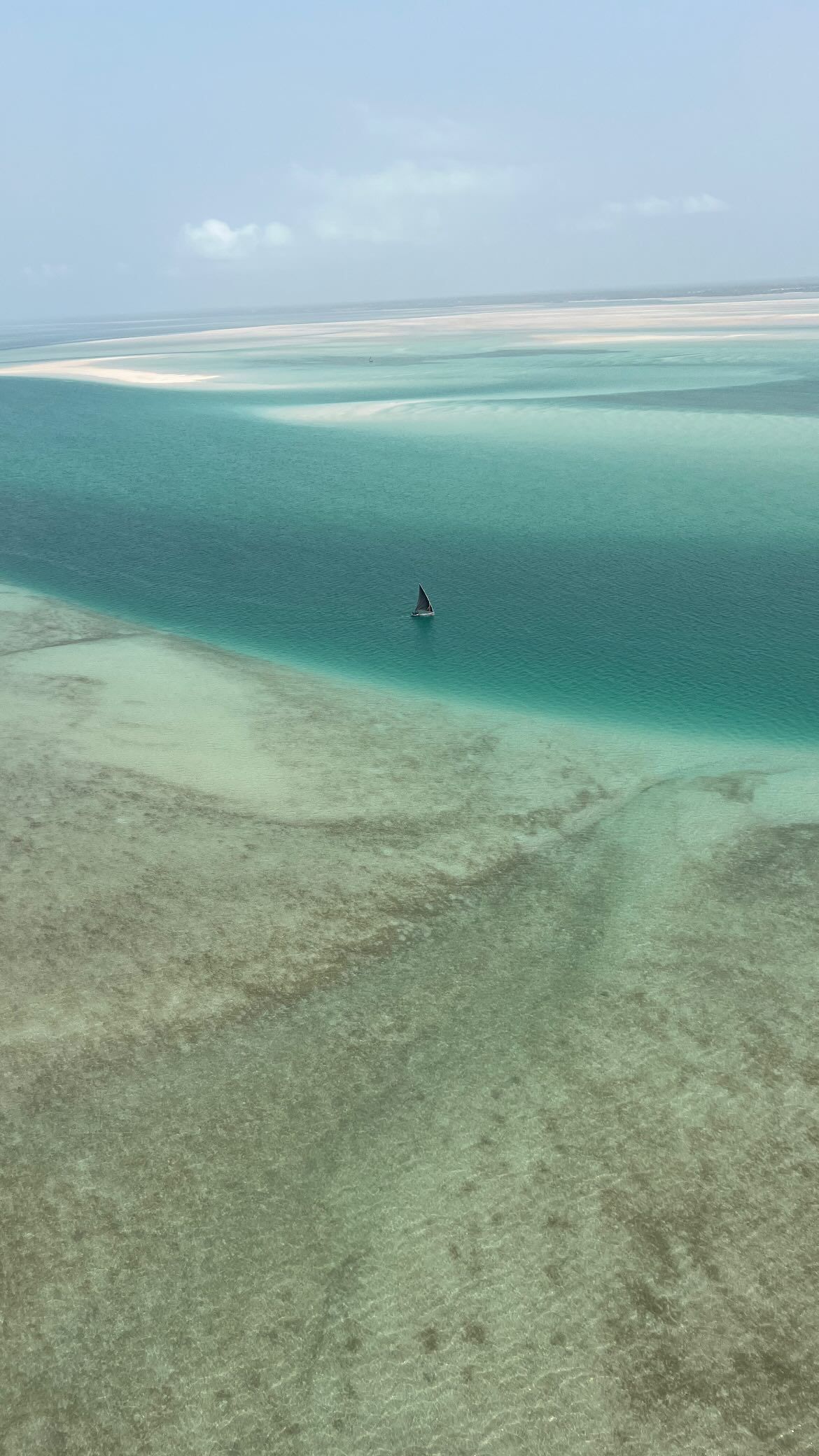 Vista aerea di una singola barca a vela su vaste acque turchesi e poco profonde con banchi di sabbia in lontananza.