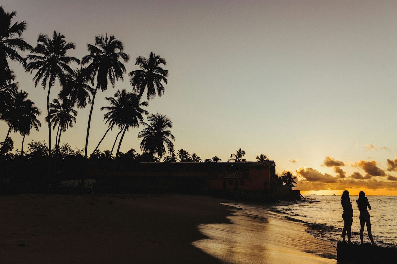 Le sagome di due persone che guardano il tramonto da una spiaggia tropicale con palme.
