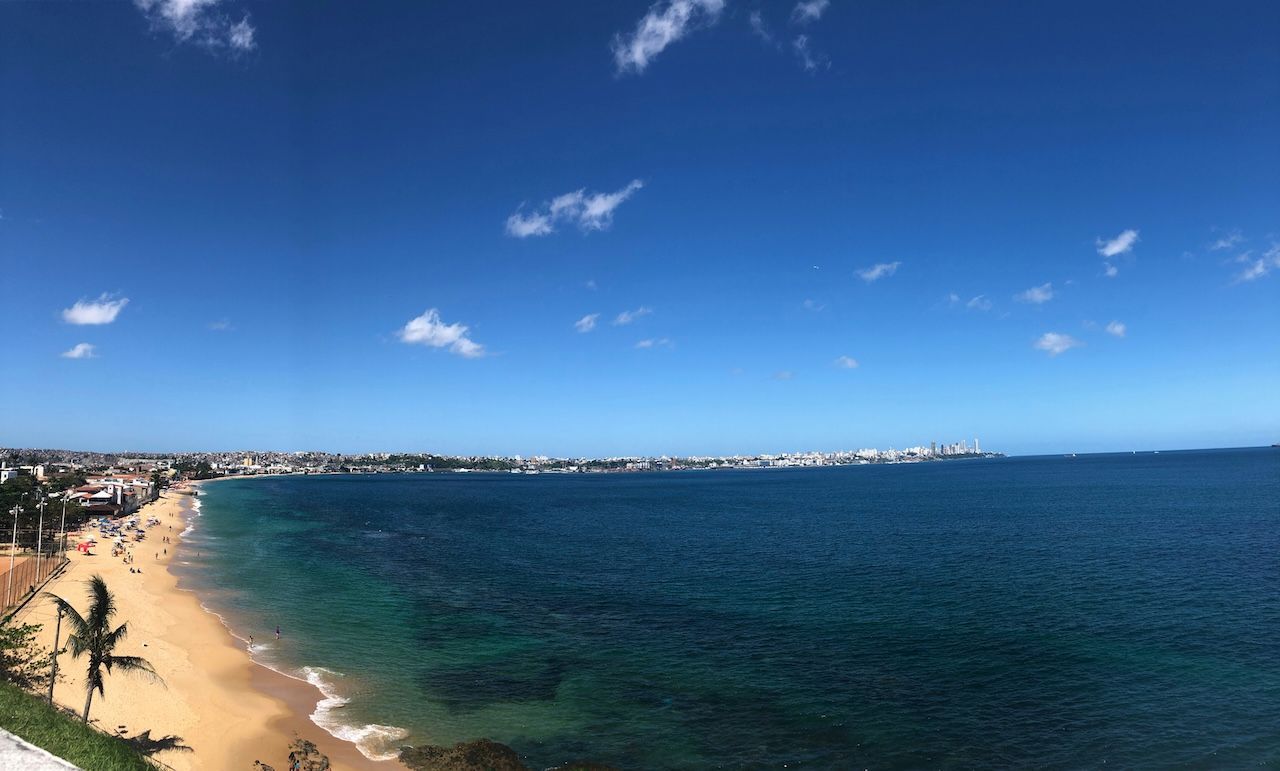 Una vista panoramica dall'alto di una costa cittadina con una lunga spiaggia di sabbia, acque turchesi e un cielo azzurro limpido con qualche nuvola.