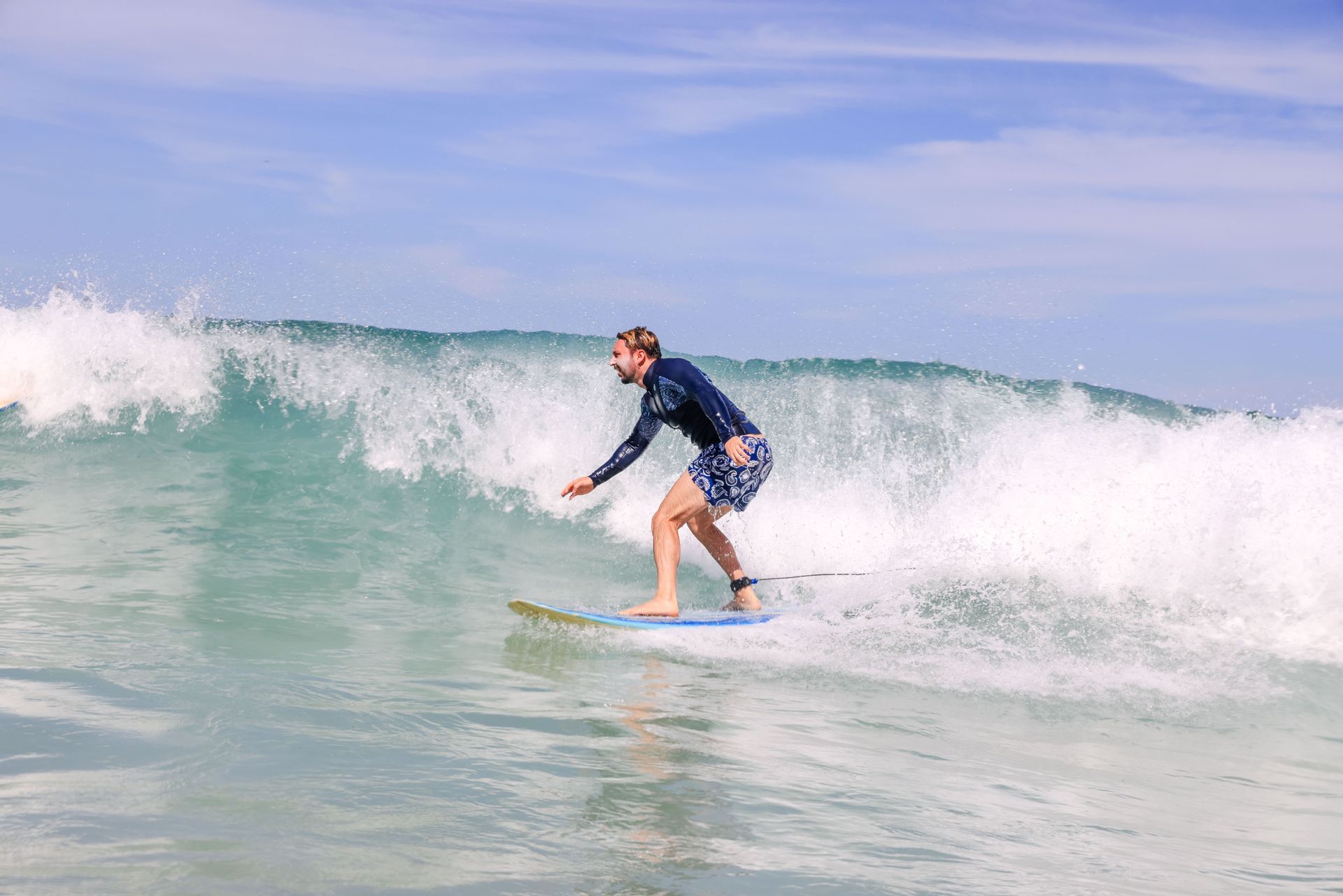 Un uomo con una rash guard e pantaloncini si equilibra su una tavola da surf mentre cavalca un'onda dell'oceano sotto un cielo parzialmente nuvoloso.