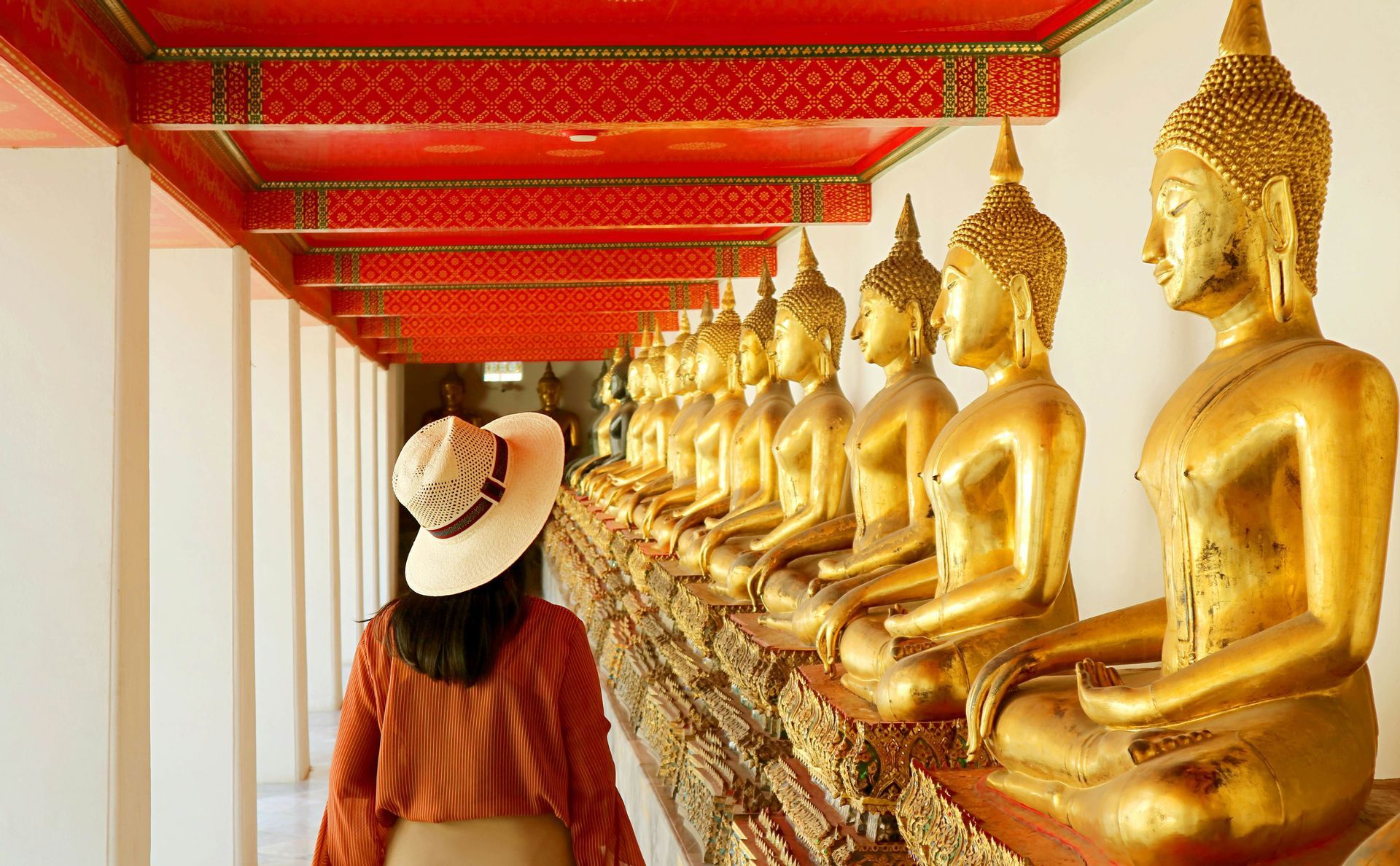 A woman wearing a straw hat walks through a temple corridor lined with a long row of golden Buddha statues.
