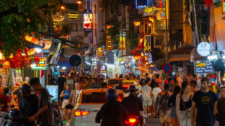 Una grande folla di persone cammina lungo una stretta via di città di notte, piena di auto, moto e luminose insegne al neon colorate.
