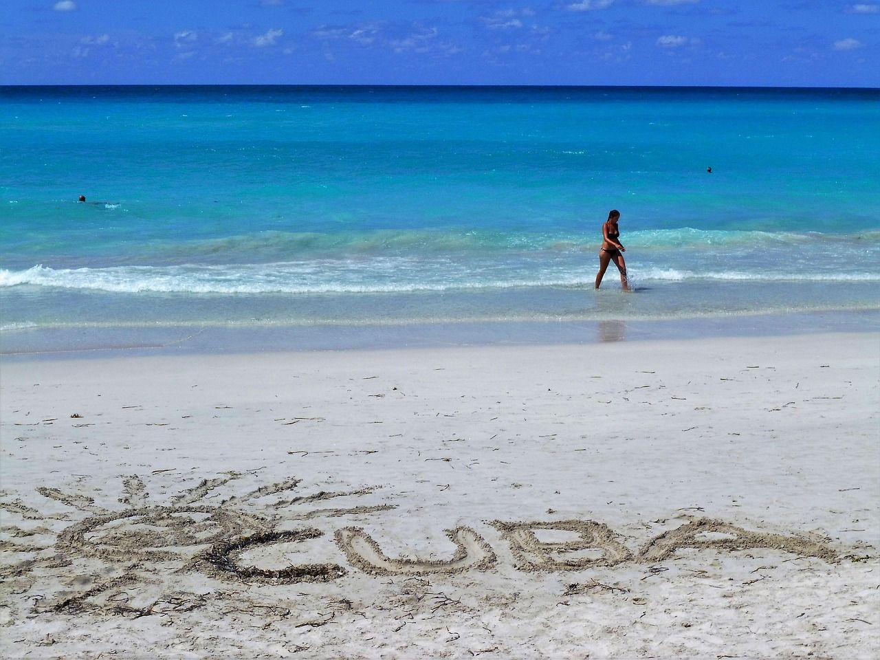 La parola 'CUBA' e un sole sono disegnati nella sabbia bianca di una spiaggia mentre una persona cammina nell'oceano turchese.