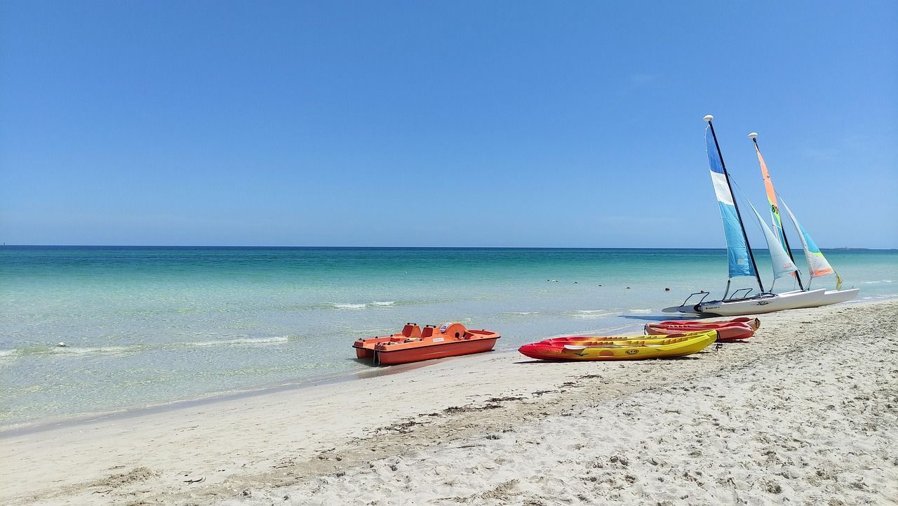 Catamarani, kayak e un pedalò sono adagiati sulla sabbia accanto all'acqua turchese sotto un cielo azzurro limpido.